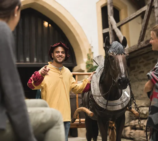 Rievocatore medievale guida visitatori accanto a un cavallo di legno in un cortile storico