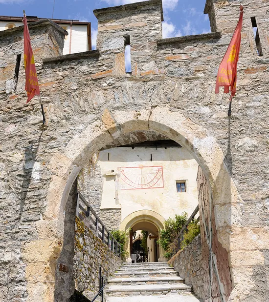 Porta in pietra e scale verso castello medievale con bandiere rosse e meridiana sopra l'arco