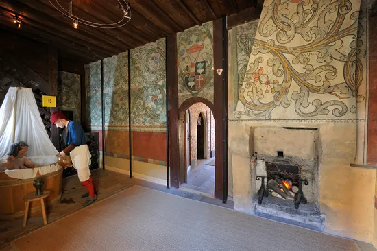 Historischer Raum mit mittelalterlicher Badeszene, Wandfresken und brennendem Kamin