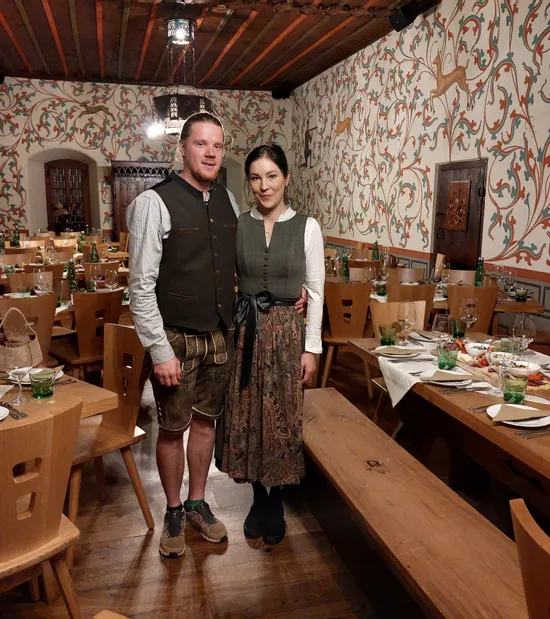 Couple in traditional Bavarian clothing standing in decorated dining hall set for dinner