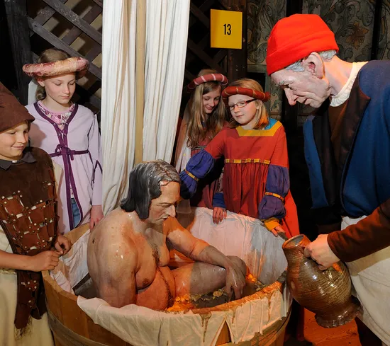 Mittelalterliche Szene mit Menschen in historischen Kostümen um einen Mann in einer Holzzuber-Badewanne