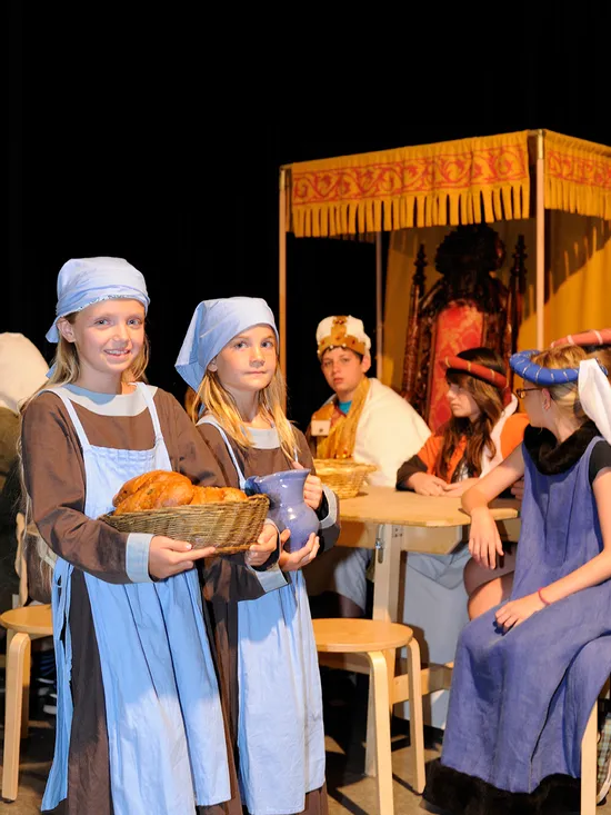 Kinder in mittelalterlichen Kostümen bei einem Theaterstück mit Essensrequisiten und Thron im Hintergrund
