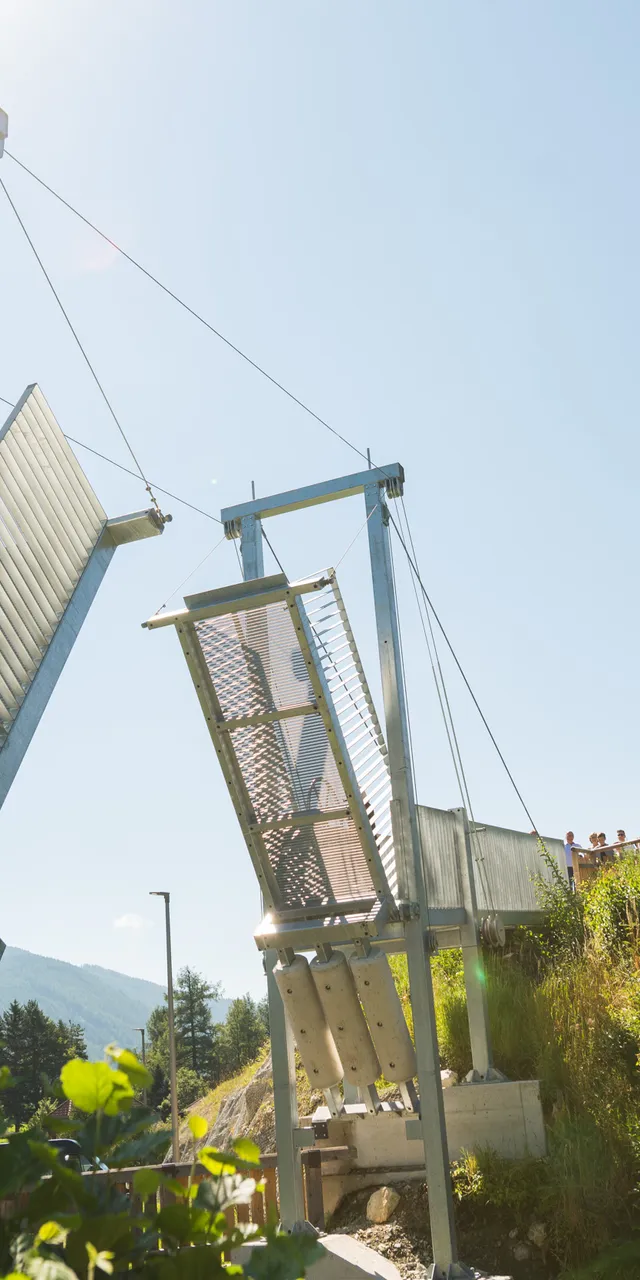 Moderne Hängebrücke mit offenen Metallplatten nahe einer historischen Steinfestung an einem sonnigen Tag