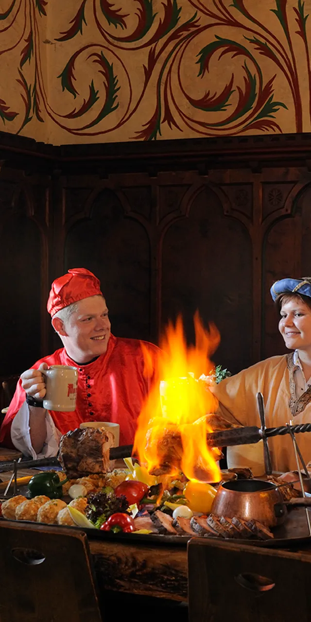 Menschen in mittelalterlichen Kostümen bei einem Festmahl an einem Holztisch mit Feuerbraten