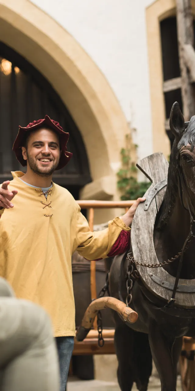 Mittelalter-Darsteller führt Besucher neben hölzernem Pferd in historischem Innenhof