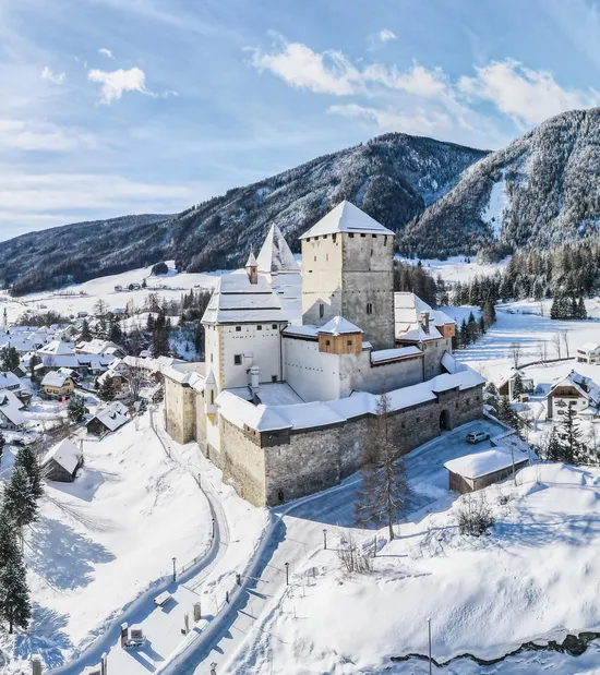 Verschneites Alpendorf mit mittelalterlicher Burg und bewaldeten Bergen im Wintersonnenlicht