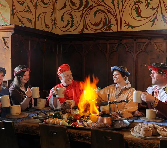 Group in medieval costumes enjoying feast with fire-roasted meat and drinks in rustic banquet hall