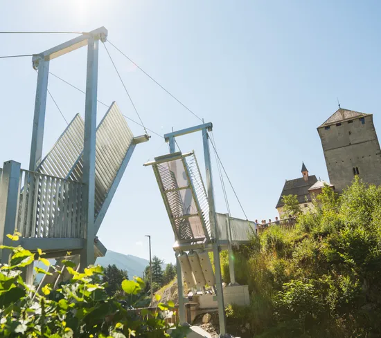 Moderne Hängebrücke mit offenen Metallplatten nahe einer historischen Steinfestung an einem sonnigen Tag