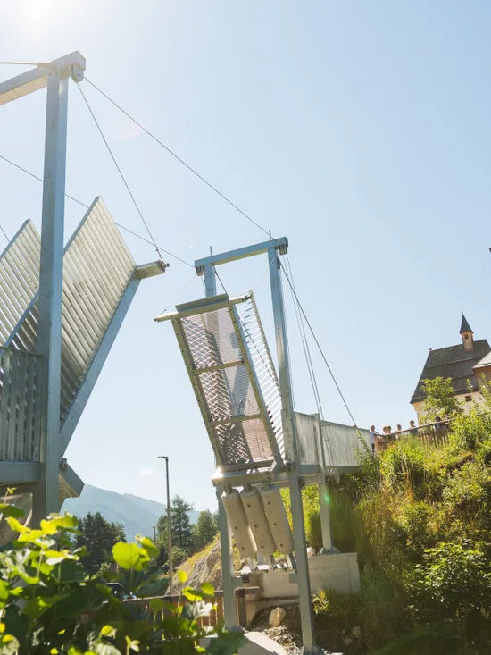 Moderne Hängebrücke mit offenen Metallplatten nahe einer historischen Steinfestung an einem sonnigen Tag